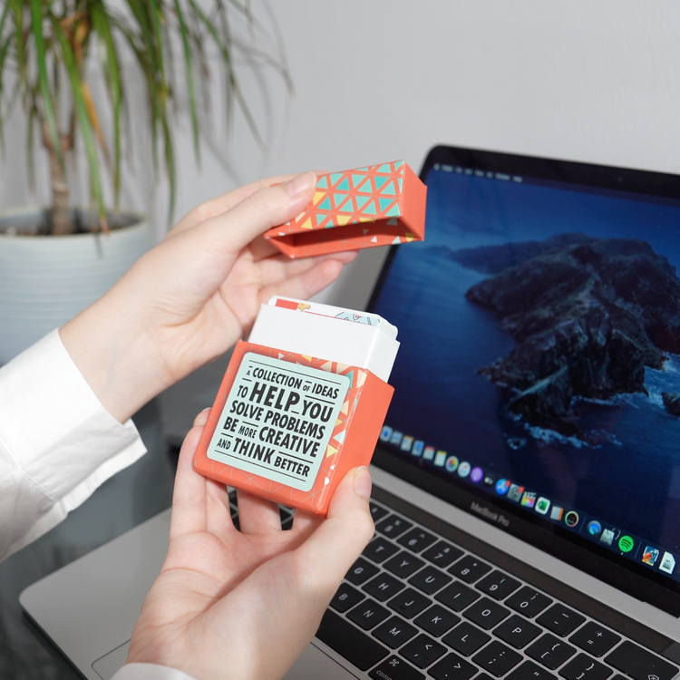 A person holds The Unstuck Box of career coaching cards in front of a laptop, showcasing a deck designed to enhance strategic thinking and problem-solving skills.